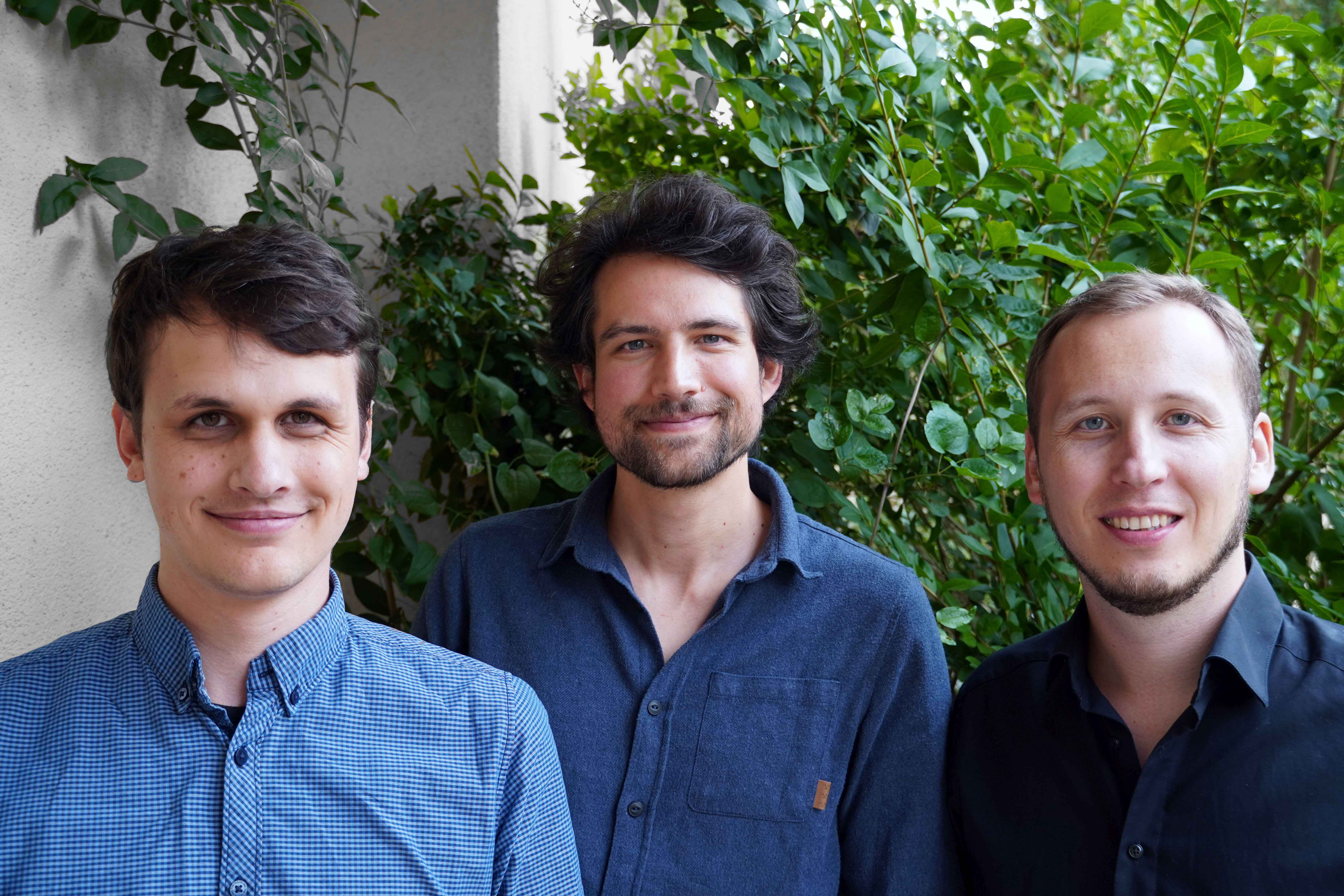 Three young men can be seen in front of a green façade, smiling friendly into the camera.